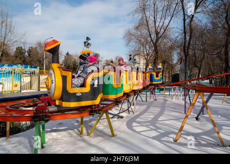 Kinder und ihre Großmutter fahren auf hohen Schienen mit einem Kinderzug über Schneewehen zwischen Bäumen in einem städtischen Vergnügungspark. Stockfoto