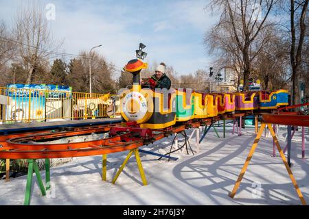 Eine Frau mit einem Handy in den Händen fährt auf hohen Schienen über Schneewehen zwischen Bäumen in einem städtischen Vergnügungspark einen Kinderzug. Stockfoto