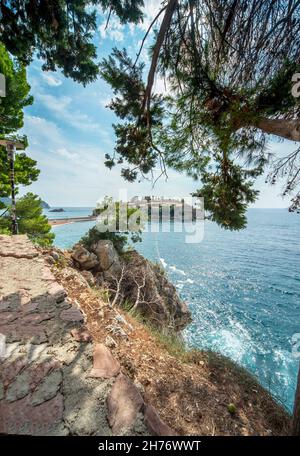 Eine exklusive, private, idyllische Insel, ein Hotel-Dorf genannt, die ins Meer hinausragt, von einem malerischen Pfad gesehen, der sich entlang eines felsigen bewaldeten Stranges schlängelt Stockfoto