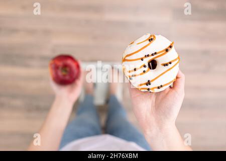 Eine Frau steht auf der Waage, hält einen Donut und einen Apfel in den Händen, Draufsicht. Stockfoto
