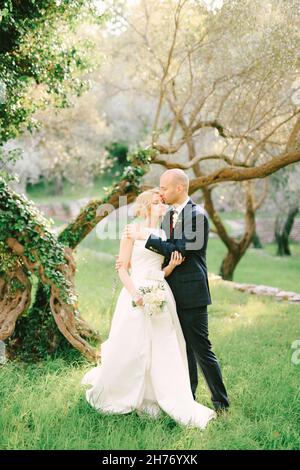 Bräutigam in einem blauen Anzug umarmt und küsst auf der Stirn Braut in einem weißen Kleid mit einem Blumenstrauß, der im Park vor dem Hintergrund eines steht Stockfoto