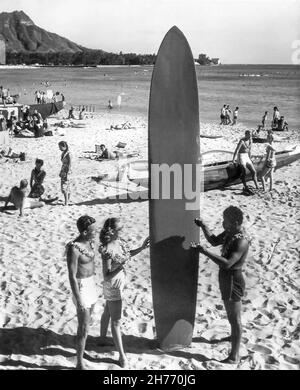 Vintage Waikiki Beach, Hawaii, Reisefoto von den 1960s Touristen mit einem hawaiianischen Surfer, der ein hölzernes Longboard hält, mit einem aufgestacheltem Outrigger-Kanu und Diamond Head im Hintergrund. (USA) Stockfoto