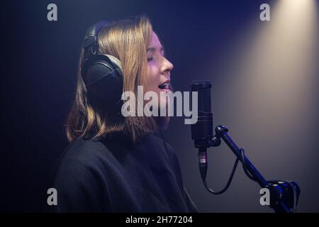 Eine niedliche junge Frau mit Kopfhörern singt in einem dunklen Raum in ein Mikrofon. Stockfoto