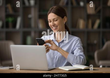 Glückliche Geschäftsfrau sitzt am Arbeitsplatz in der Nähe des Laptops und hält das Smartphone Stockfoto