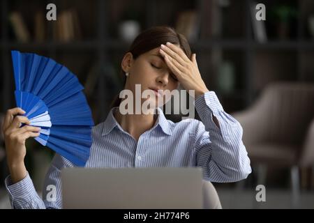 Ungesunde überhitzte Frau sitzt am Arbeitsplatz und winkt mit Handventilator Stockfoto