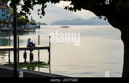 Blick auf den ruhigen Zugersee in der Schweizer Stadt Zug mit einem kleinen Holzhafen und einem jungen Mädchen, das bei Sonnenaufgang über den See blickt. Stockfoto