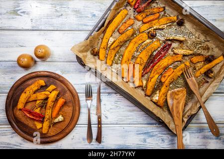 Gerösteter Kürbis hokkaido und Gemüse auf Holzplatte. Gebackenes Gemüse als herbstliches Abendessen. Stellen Sie den Esstisch mit rustikalem Besteck Stockfoto