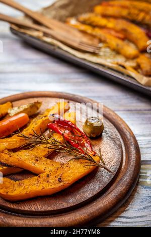 Gebackener Kürbis hokkaido und gebratenes Gemüse auf Holzplatte. Backblech auf weißem Tisch. Vegetarische Gerichte Stockfoto