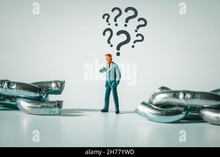 Makrofoto einer durchdachten Geschäftsmann-Figur, die zwischen einer zerbrochenen Kette steht. Fragezeichen über dem nachdenkenden Miniaturmenschen. Konflikt, Konkurs, Stockfoto