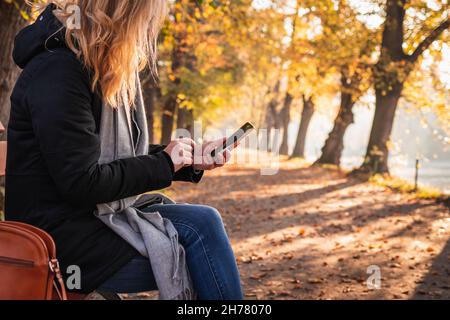 Frau, die im Herbstpark auf der Bank sitzt und Smartphone benutzt. Entspannung und SMS auf dem Handy während der Städtereise im Herbst Stockfoto
