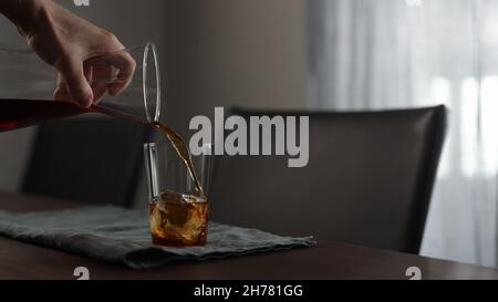 Mann gießt frischen Kaffee über Eisfelsen in Glas auf Holztisch, breites Foto Stockfoto