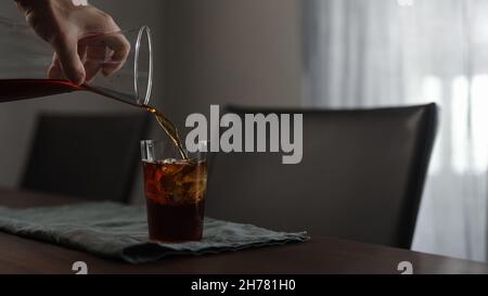 Mann gießt frischen Kaffee über Eisfelsen in Glas auf Holztisch, breites Foto Stockfoto