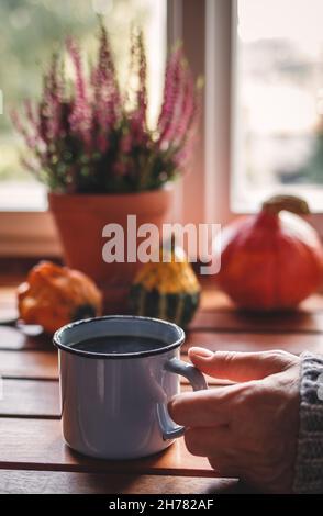 Genießen Sie starken schwarzen Kaffee. Frau hält Tasse Heißgetränk in gemütlichen Innenraum mit Herbstdeko. Sich zu Hause ruhig und wohl fühlen Stockfoto