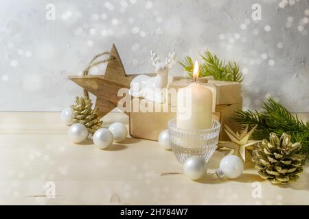 Weihnachtsdekoration mit einer warmweißen Kerze, Porzellanhirschen, Kugeln, Holzsternen und Tannenzweigen auf hellgrauem Hintergrund mit schneebedecktem Bokeh Stockfoto