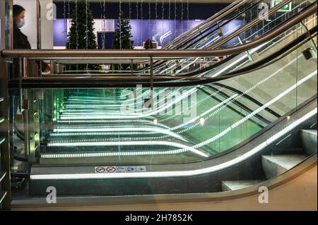Abstrakte Rolltreppen in einem Kaufhaus mit einem unscharfen, nicht erkennbaren Kunden im Profil, der eine Gesichtsmaske trägt, Konzept für Wirtschaft und die Wirkung von Stockfoto