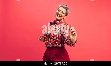 Ich will nur tanzen. Unbeschwerte junge Frau, die vor einem roten Hintergrund tanzt und fröhlich lächelt. Junge Frau mit gefärbten Haaren hat Spaß Stockfoto