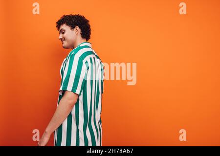 Fröhlicher junger queerer Mann, der vor einem orangefarbenen Hintergrund steht. Seitenansicht eines glücklichen jungen Generation z Hipsters, der selbstbewusst im Stehen aussieht Stockfoto
