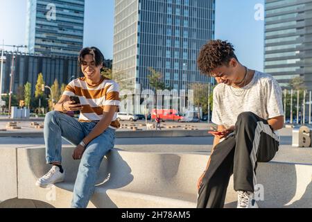 Zwei glückliche Teenager-Freunde, jeder von ihnen schaute auf ihr Telefon in einer Bank auf der Straße. Stockfoto