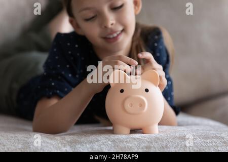 Kleines Mädchen, das auf dem Sofa liegt, steckte eine Münze in das Sparschwein Stockfoto