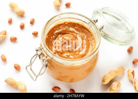 Erdnussbutter in einem Glas und verstreute Erdnüsse isoliert auf weißem Hintergrund. Stockfoto