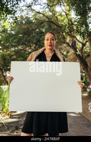 Selbstbewusste, schicke Frau, die die Kamera anschaut und dabei ein weißes Plakat hochhält. Frau mittleren Alters, die ein leeres Banner zeigt. Weibliche Aktivistin, die al steht Stockfoto