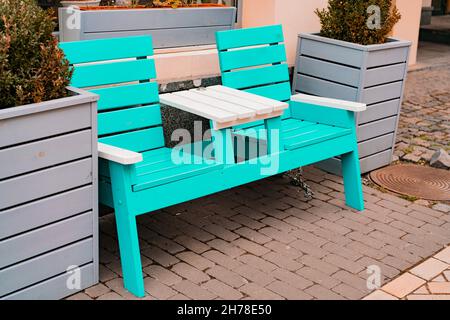 Türkisfarbene Bank mit kleinem weißen Tisch auf der Straße. Graue Holztöpfe. Im Freien. Stadt. Restaurant. Gehweg. Leer. Keine Personen. Dekoration Stockfoto