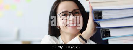 Junge Frau mit Brille, die viele Ordner mit Dokumenten im Büro hält Stockfoto