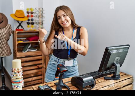 Die junge Brünette hält ein Banner mit offenem Text im Einzelhandelsgeschäft, das mit der Hand und den Daumen nach hinten zeigt, und lächelt zuversichtlich Stockfoto