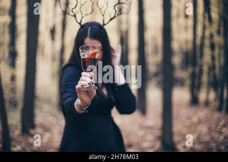 Geheimnisvolle Frau in schwarzen Kleidern, die ein Stirnband mit gotischen Hirschgeweihen trägt und Glühwein in der Hand aushält, während sie während des Hal allein im Herbstwald spazieren geht Stockfoto