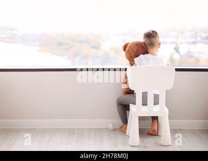 Rückansicht eines kleinen Jungen mit Teddybär, der auf einem Stuhl sitzt und aus dem Fenster auf die herbstliche Stadtlandschaft blickt, während er zu Hause auf die Mutter wartet. Konzept für die Kindheit Stockfoto