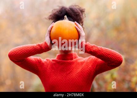 Junge Mischrassen-Frau mit lockigen Haaren, die orangen Kürbis hält, während sie vor einem verschwommenen Herbstwald steht. Herbstsaison und Erntekonzept Stockfoto