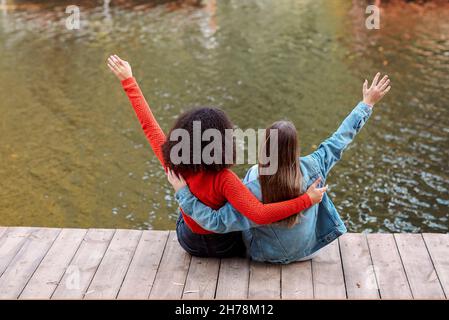 Rückansicht von zwei glücklichen multirassischen Freundinnen, die in der Nähe von River sitzen und mit Glück die Hände in der Luft heben, zwei besten Freundinnen verschiedener Rassen Stockfoto