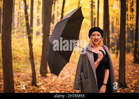 Schöne junge stilvolle Frau trägt Baskenmütze und Mantel mit Regenschirm beim Spaziergang im Herbstwald am regnerischen Tag, glückliche Hündin mit lila Lippenstift h Stockfoto