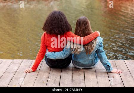 Rückansicht von zwei glücklichen multirassischen Freundinnen, die in der Nähe des Flusses mit Glück sitzen, zwei besten Freundinnen verschiedener Rassen, die sich in der Nähe des Teiches auf der Haut entspannen Stockfoto