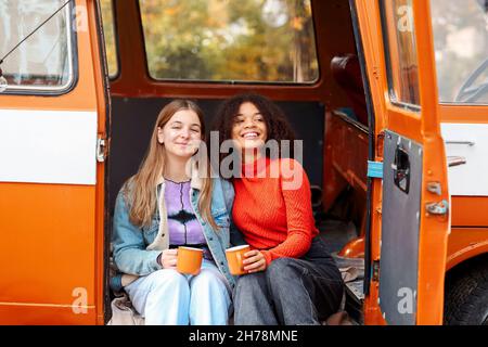 Zwei glückliche multirassische Freundinnen sitzen im Freien, trinken heißen Tee und reden bei Treffen im Park am Herbsttag, lächelnd weibliche beste Freunde genießen Stockfoto