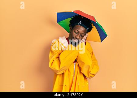 Junger afroamerikanischer Mann mit gelbem Regenmantel, müde schlafend, träumend und mit Händen zusammen posiert, während er mit geschlossenen Augen lächelt. Stockfoto