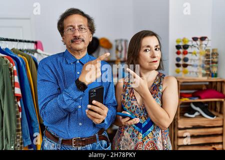 Interrassisches Paar im mittleren Alter im Einzelhandelsgeschäft mit Smartphone, das mit dem Handfinger auf die Seite zeigt und Werbung, ernstes und ruhiges Gesicht zeigt Stockfoto