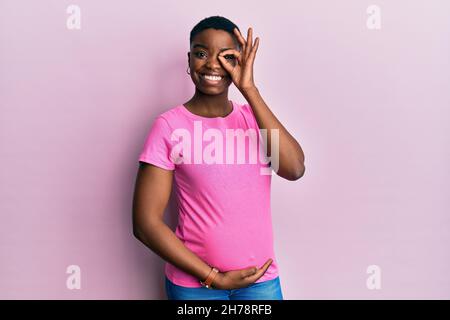 Junge afroamerikanische Frau erwartet ein Baby, berühren schwanger Bauch tun ok Geste mit Hand lächelnd, Auge durch die Finger mit glücklichen Gesicht schauen Stockfoto