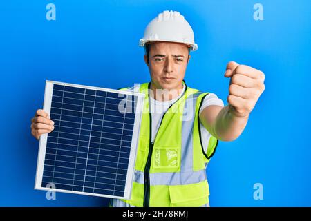 Hübscher junger Mann, der eine Photovoltaikanlage hält, ist verärgert und frustriert, vor Wut schreiend, verrückt vor Wut und erhobener Hand Stockfoto