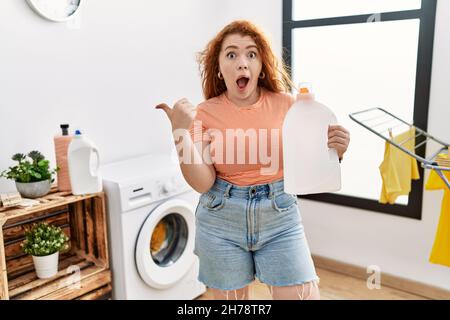 Junge Rothaarige Frau, die Wäsche waschen und die Waschmittelflasche mit dem Daumen nach oben zur Seite zeigt, lächelt glücklich mit offenem Mund Stockfoto