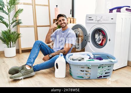 Junger hispanischer Mann, der schmutzige Wäsche in die Waschmaschine legt, lächelt und zeigt mit einem Finger auf den Kopf, großartige Idee oder Gedanke, gute Erinnerung Stockfoto