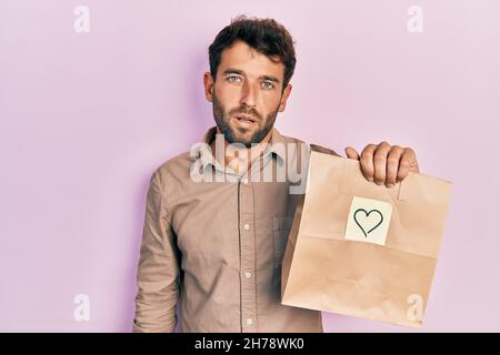 Schöner Mann mit Bart halten Lieferung Papiertasche mit Herz Erinnerung suchen müde und müde, erschöpft für Müdigkeit und Kater, faule Augen in der Stockfoto