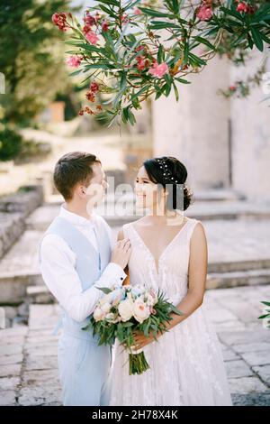 Braut und Bräutigam umarmen sich unter einem blühenden Oleander, die Braut hält einen Strauß in ihren Händen Stockfoto