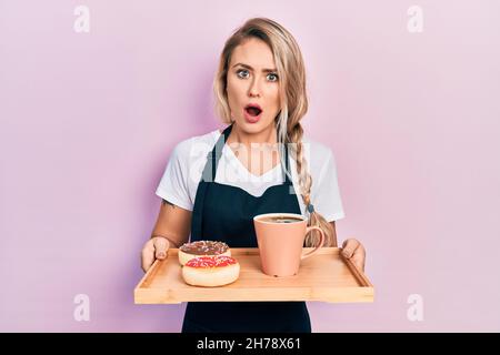 Schöne junge blonde Frau mit Kellnerin Schürze hält Frühstückstablett im Schockgesicht, sieht skeptisch und sarkastisch, überrascht mit offenem Mund Stockfoto