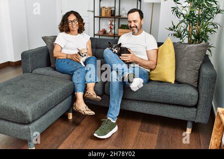 hispanische Paare mittleren Alters lächeln glücklich auf dem Sofa mit Hunden zu Hause sitzen. Stockfoto