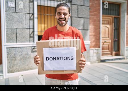 Junger hispanischer Mann hält Spendenbox für Wohltätigkeitsorganisationen im Freien und lacht laut, weil lustig verrückt Witz. Stockfoto
