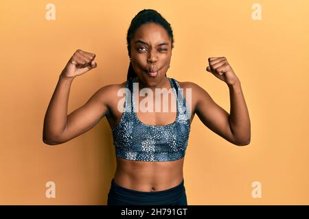 Afroamerikanische Frau mit geflochtenen Haaren, die Sportkleidung trägt und Armmuskeln zeigt, die Fische mit Mund ins Gesicht machen und Augen schielen, verrückt und komisch. Stockfoto