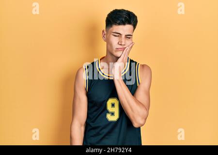 Junger hispanischer Mann trägt Basketballuniform und berührt den Mund mit der Hand mit schmerzhaftem Ausdruck wegen Zahnschmerzen oder Zahnerkrankungen an den Zähnen. Gebiss Stockfoto