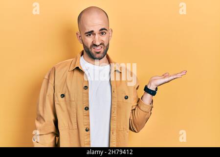 Junger kahler Mann präsentiert mit offenen Handflächen, hält etwas ahnungslos und verwirrt Ausdruck. Zweifel Konzept. Stockfoto