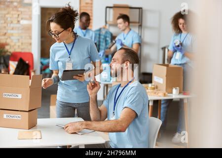 Weibliche und männliche Freiwillige arbeiten gemeinsam an einem Spendenprojekt in Innenräumen. Sie berechnen, sortieren und verpacken Artikel in Kartons Stockfoto